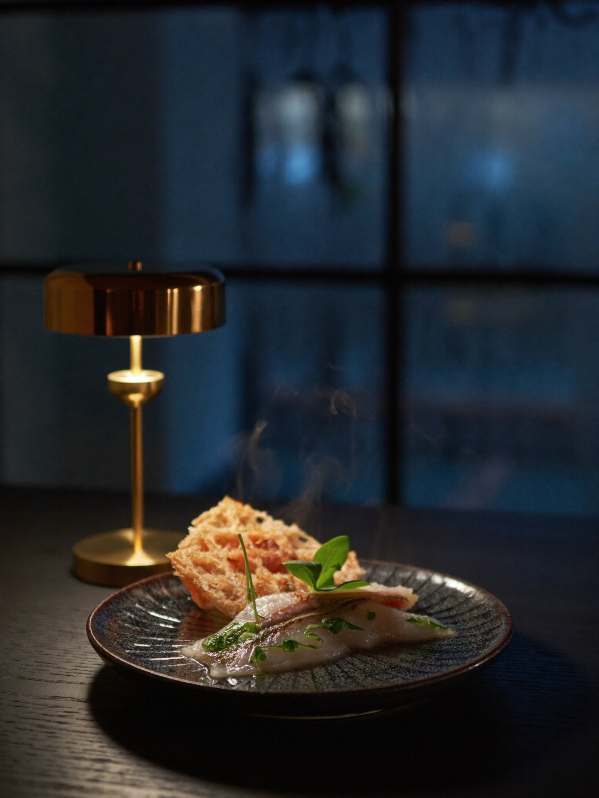 Sương — steaming modern Vietnamese dish on dark ceramic, brass lamp against a rain-blurred window